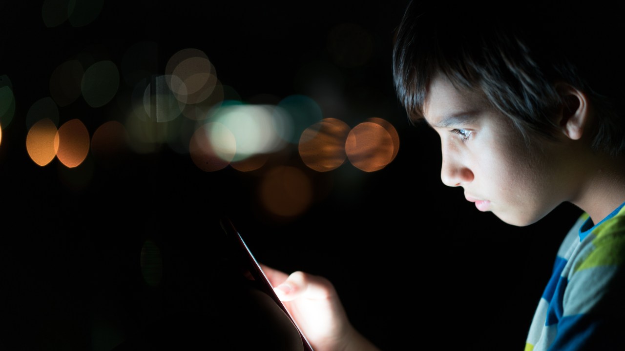 Criança usando o celular no escuro. O rosto do menino, que tem por volta de 8 anos, está iluminado pela tela.