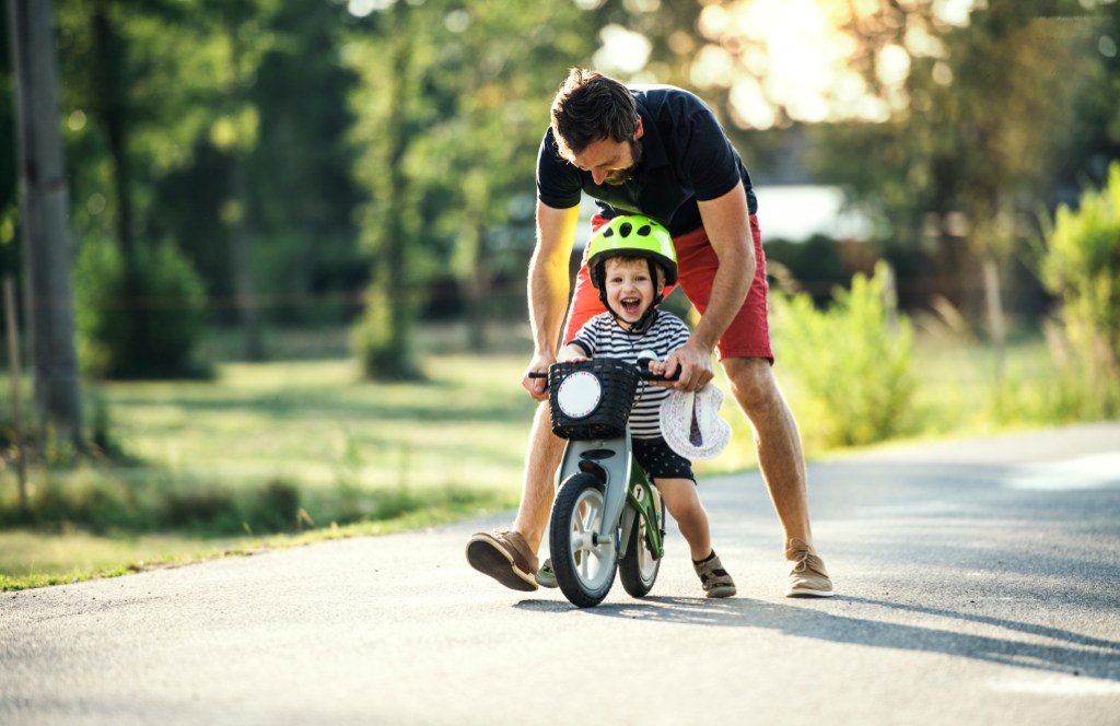 Pai ensinando o filho a andar de bicicleta na rua. O menino usa capacete verde, camiseta listrada e shorts preto com detalhes. O pai usa bermuda vermelha e camiseta preta. Eles estão sorrindo.