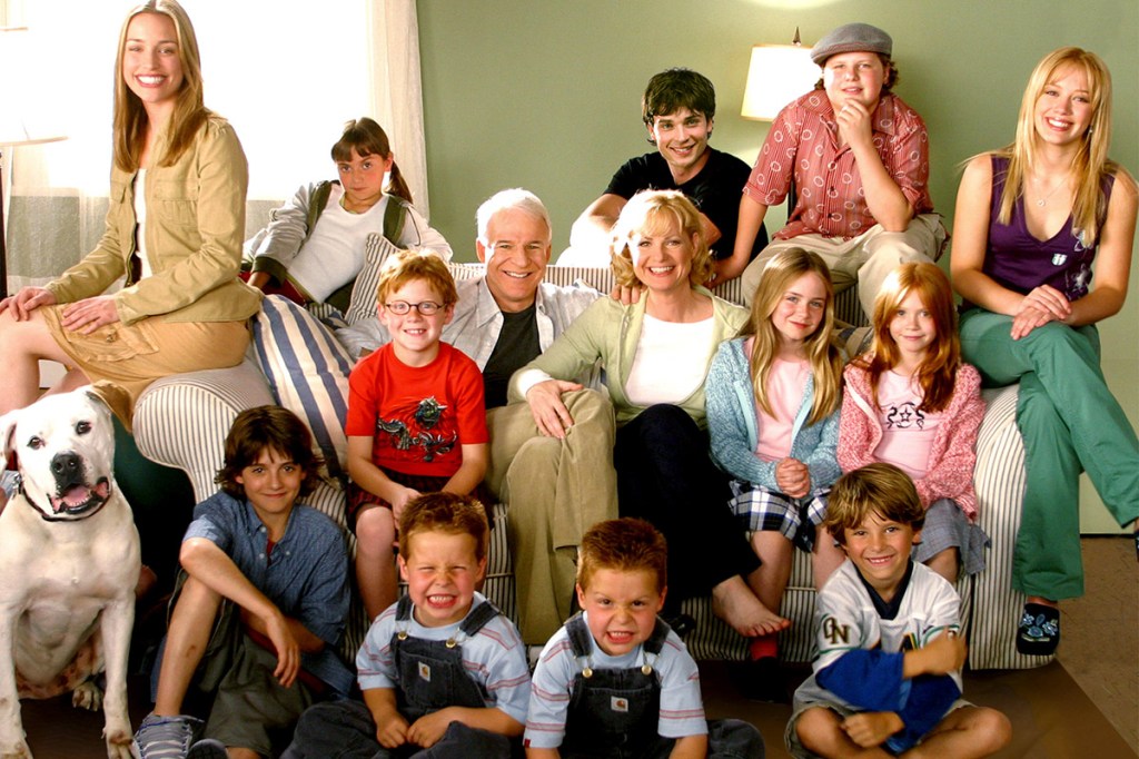 Família toda reunida na sala. Estão sentados os pais no sofá e todos os 12 filhos ao redor deles.
