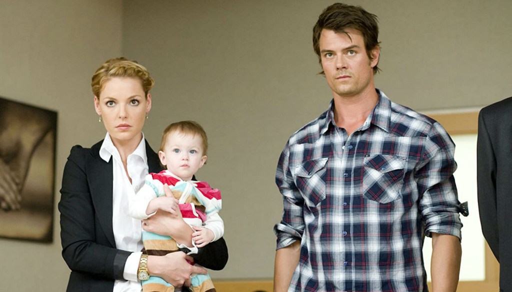Cena do filme: a personagem feminina está com a bebê no colo, o personagem masculino está ao lado dela. Estão todos olhando sérios para frente.