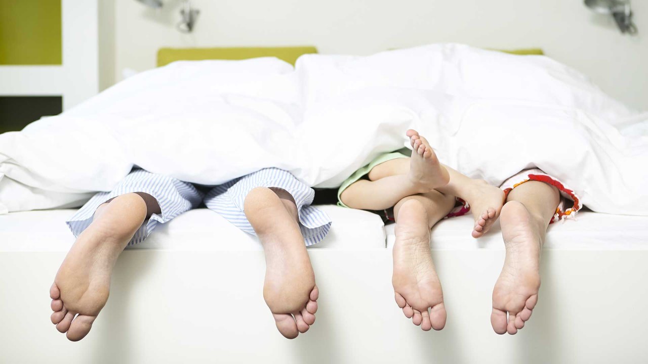 três pares de pés, de um homem, uma mulher e uma criança, em uma cama, cobertos com uma colcha branca